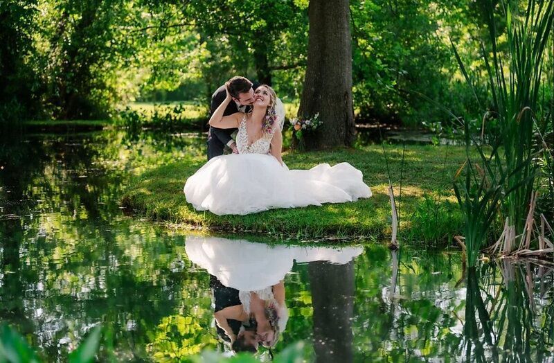 Bride and groom on grassy bank by pond; reflection in water. Couple embracing, sunny outdoor setting.