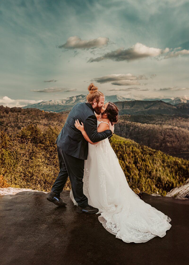 Couple embracing and kissing on a mountaintop, bride in white gown, groom in suit, scenic view.