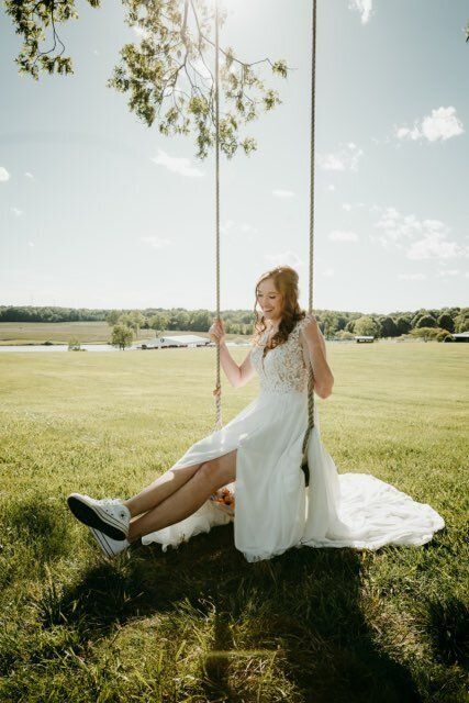 Bride in white dress on swing, sitting, smiling. Open field with trees, sunny.