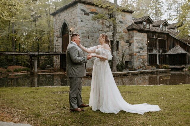 Bride touches groom's face; they stand outside a stone building. Wedding day, soft lighting.