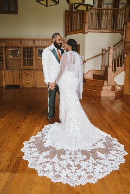 Couple in formal wear holding hands in a wood-paneled room, bride with long train, groom in white suit.