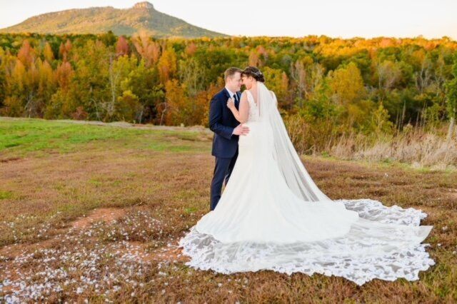 Couple embraces outdoors in wedding attire, autumn landscape with mountain backdrop.