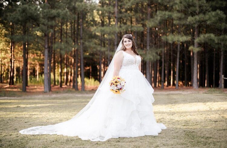 Bride in a white wedding dress holds a bouquet, standing in a field with trees in the background.