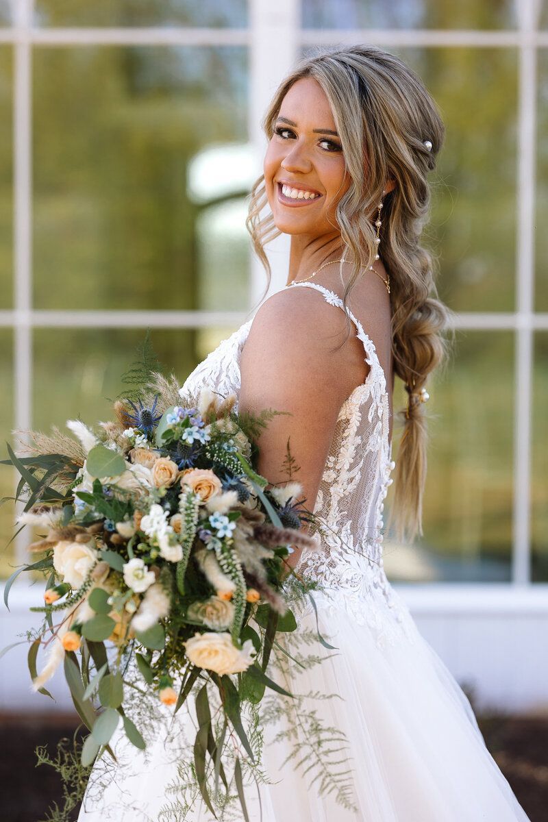 Bride in a white dress, holding a bouquet, smiling in front of a window.