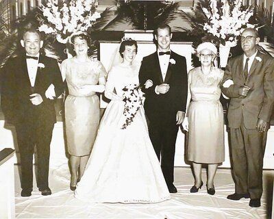 Wedding party: bride and groom stand with parents, formal attire, church setting, black and white photo.