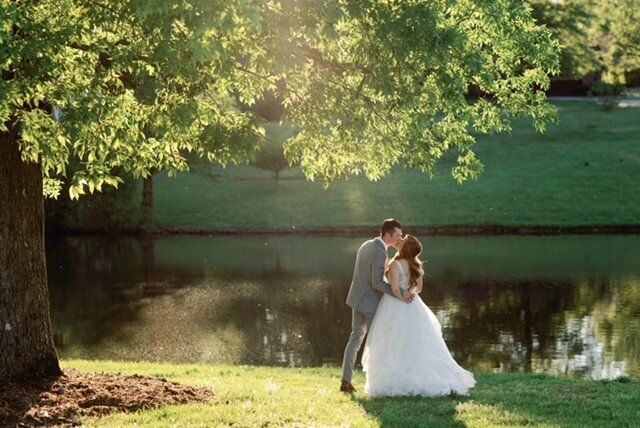 Couple kissing near a lake under a tree, backlit by sunlight. The woman wears a white dress and the man a gray suit.