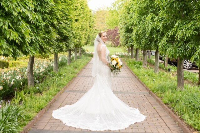 Bride in white wedding dress holding bouquet, standing on a brick path lined with trees and flowers.
