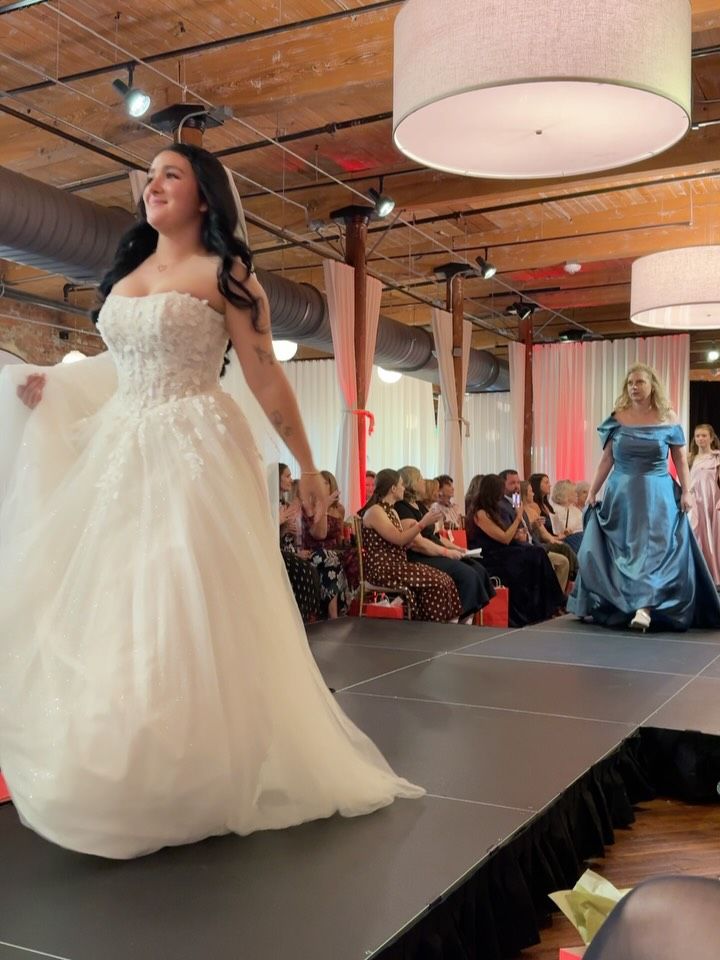 Woman in a white strapless gown on a runway, smiling. Audience seated in a red-lit venue.