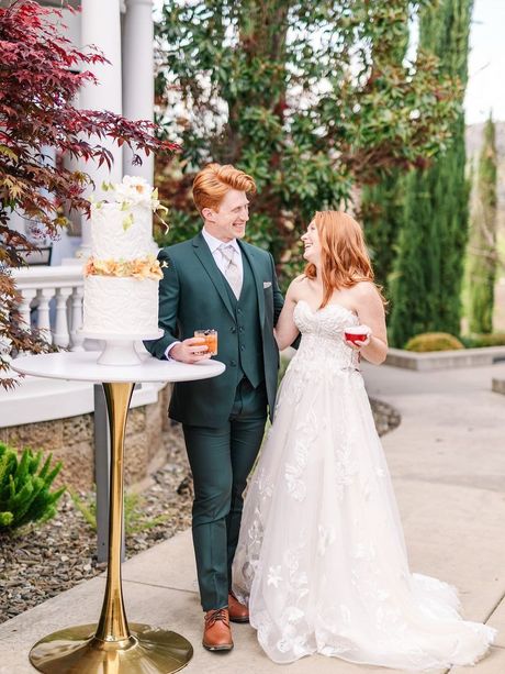 Newlyweds, a red-haired couple, laugh while standing near a tiered wedding cake outdoors. The groom wears a green suit.