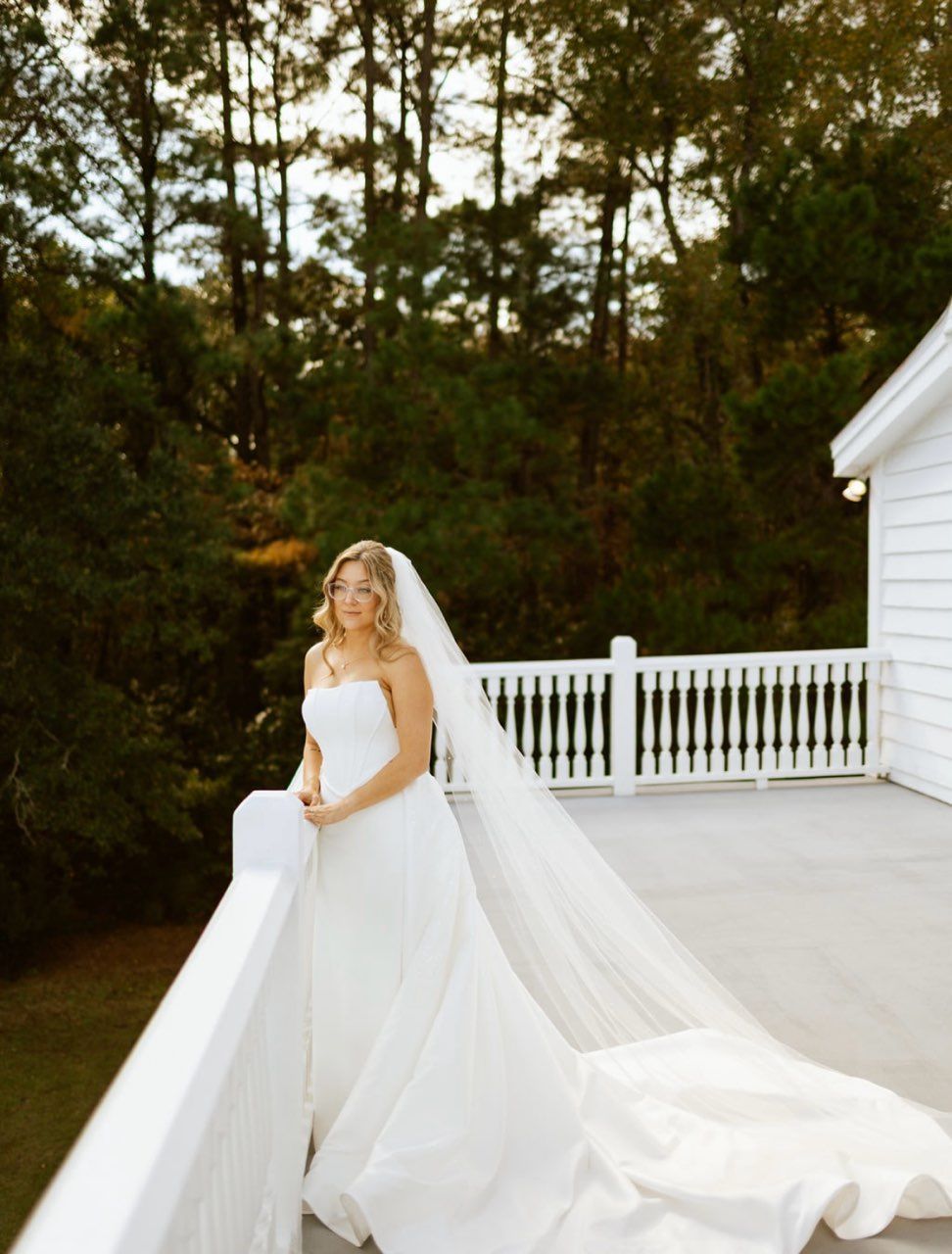 Bride in a white gown with a long veil on a white balcony with a forest in the background.