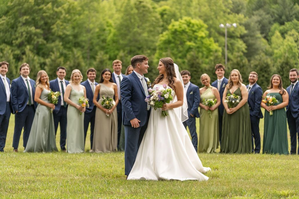 Bride and groom with wedding party; green field setting. Couple smiles, bridesmaids in green dresses, groomsmen in blue suits.
