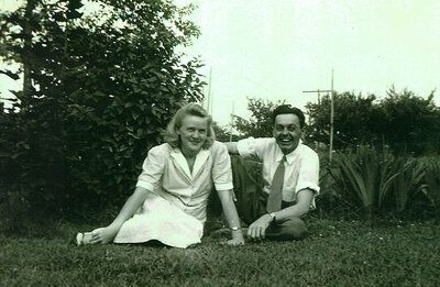 A woman and a man sit on the grass, smiling, near shrubs. They wear light-colored clothing.