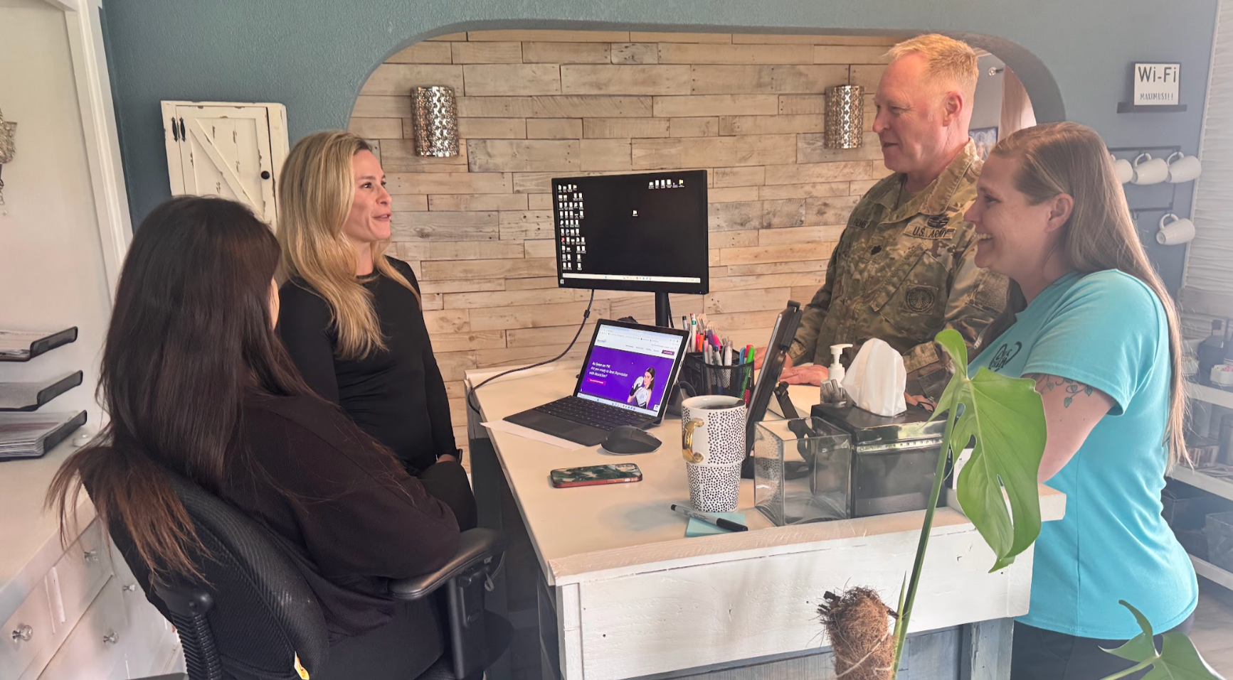 Four people in an office. A woman seated looks at a laptop while others converse around a desk with a computer and plants.