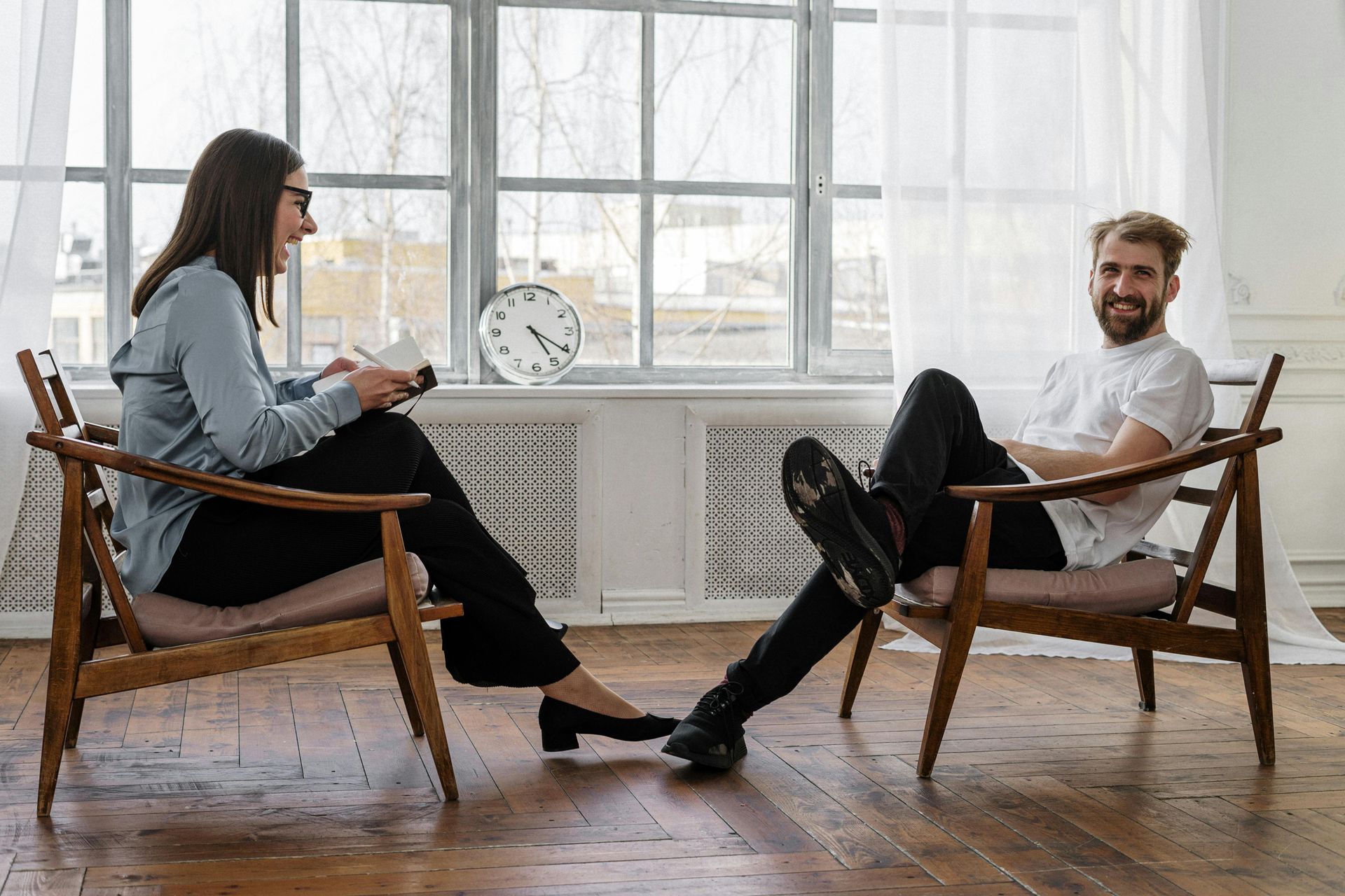 Woman reading, man smiling in armchairs facing each other by a window.
