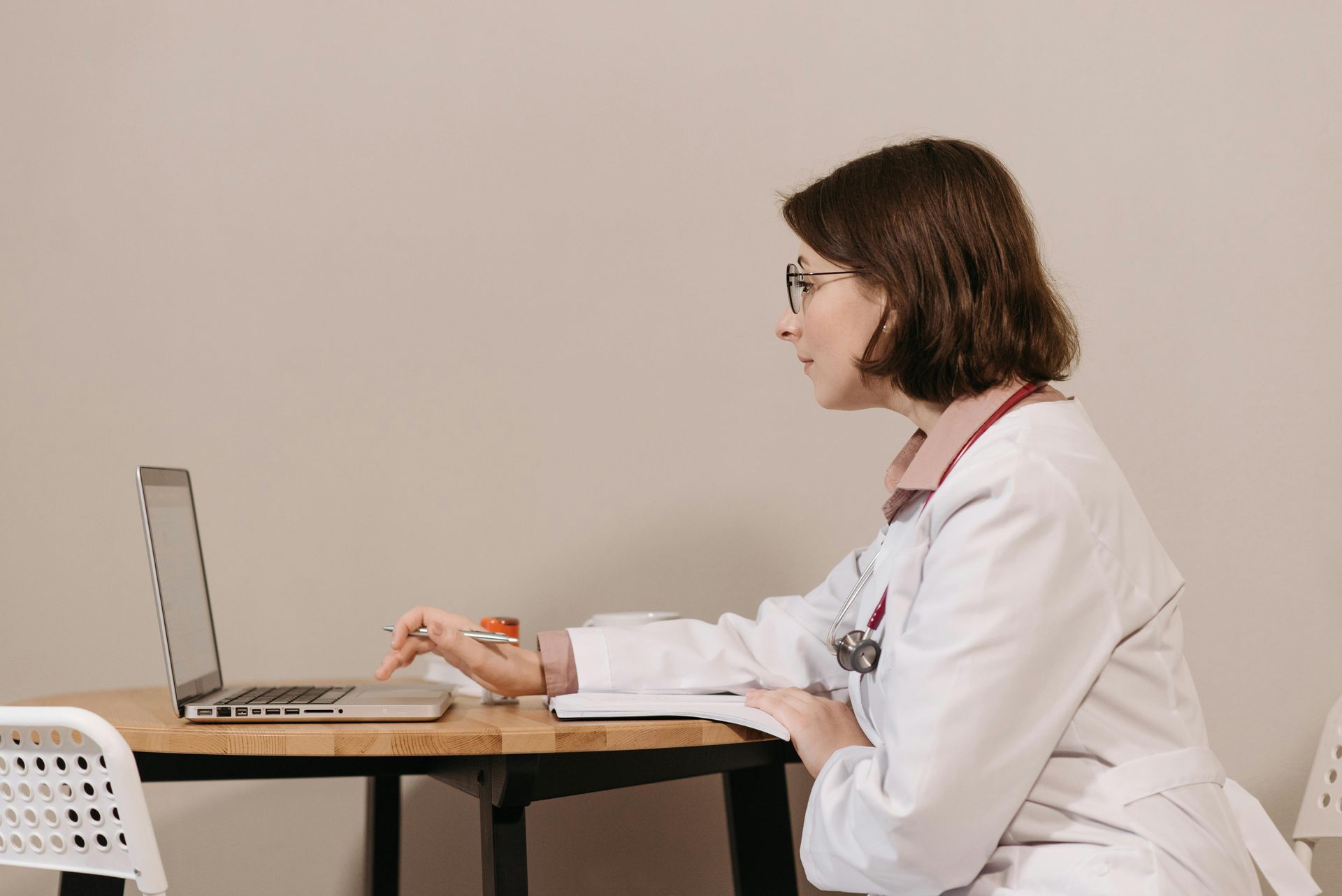 A medical professional in a lab coat sits at a small table, typing on a laptop and looking at the screen.