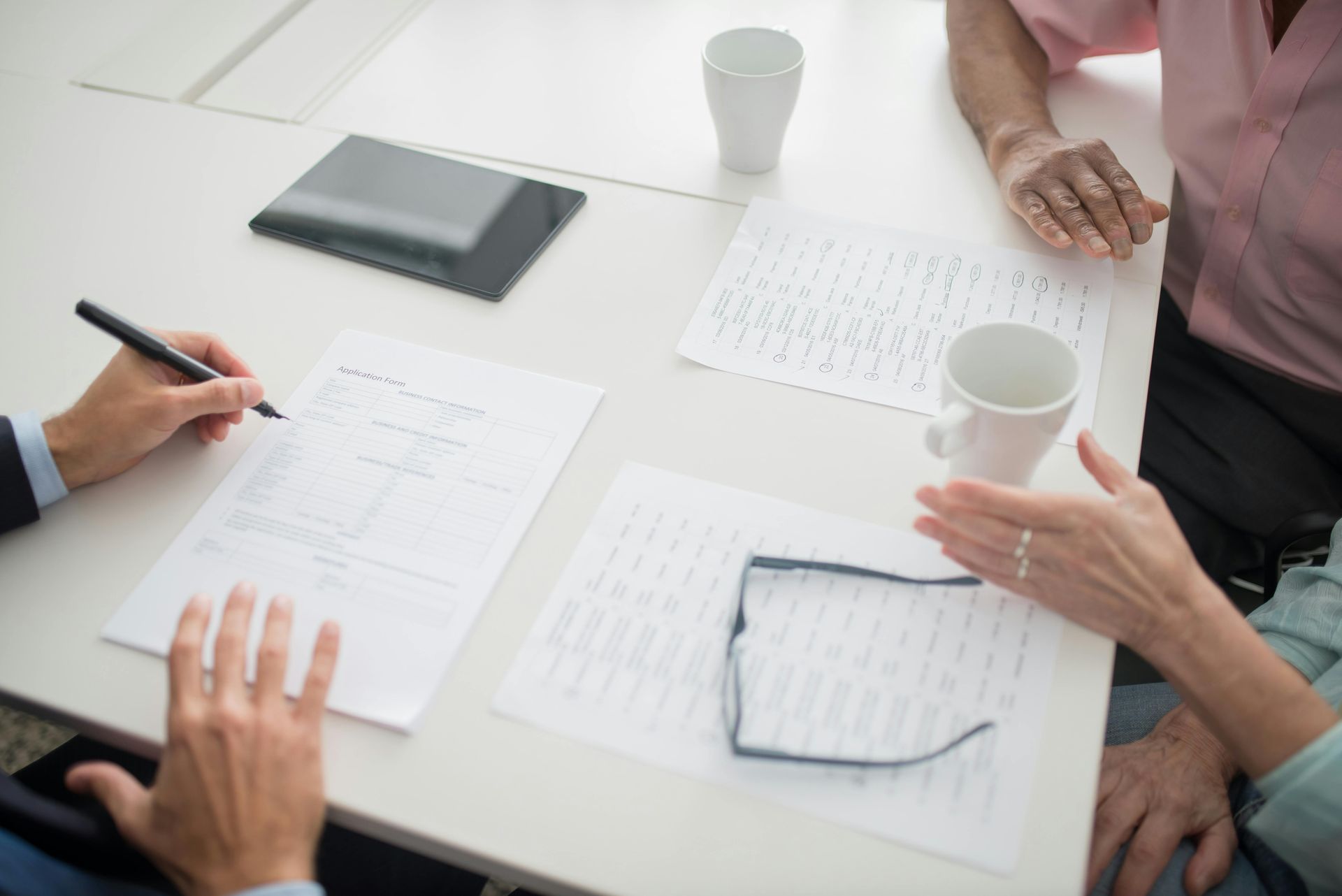 People at a table, reviewing documents and signing. A tablet and two mugs are also on the table.