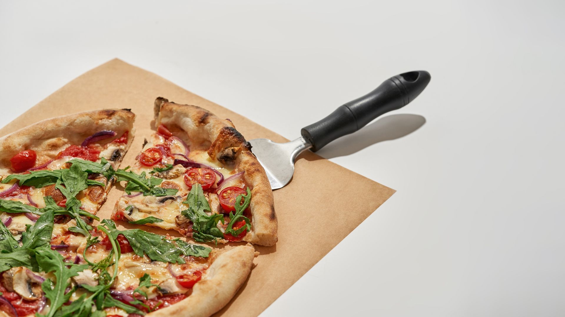 Pizza from tomato, cheese, mushroom and greens with cut slice on kitchen spatula on craft recyclable paper. Unhealthy eating. Delicious and appetizing fast food. White background in studio.