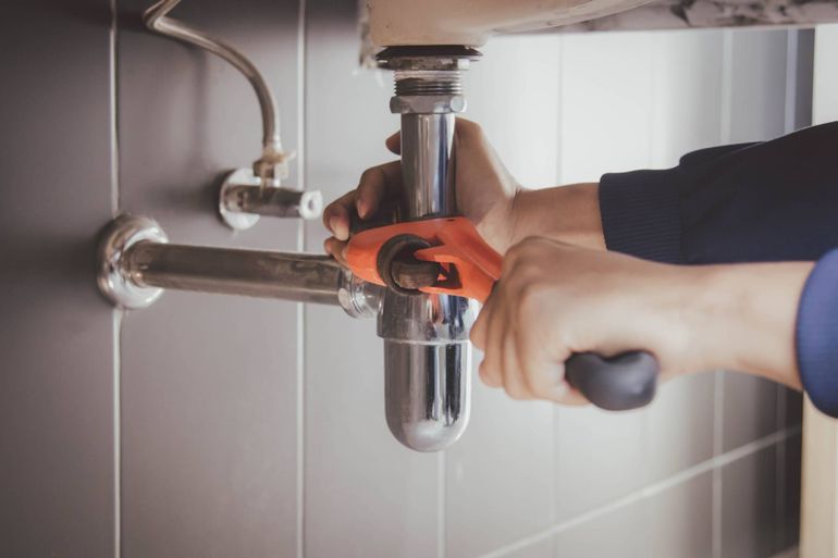 Hands tightening a chrome sink drain pipe with an orange wrench under a bathroom sink