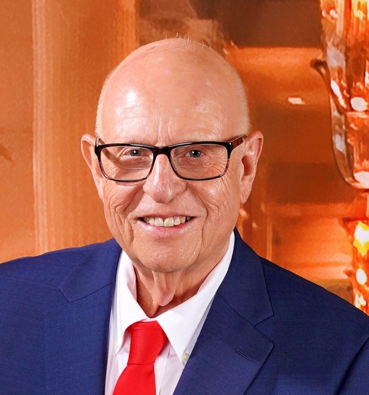 Man wearing glasses, blue suit, and red tie smiling in front of an orange background.