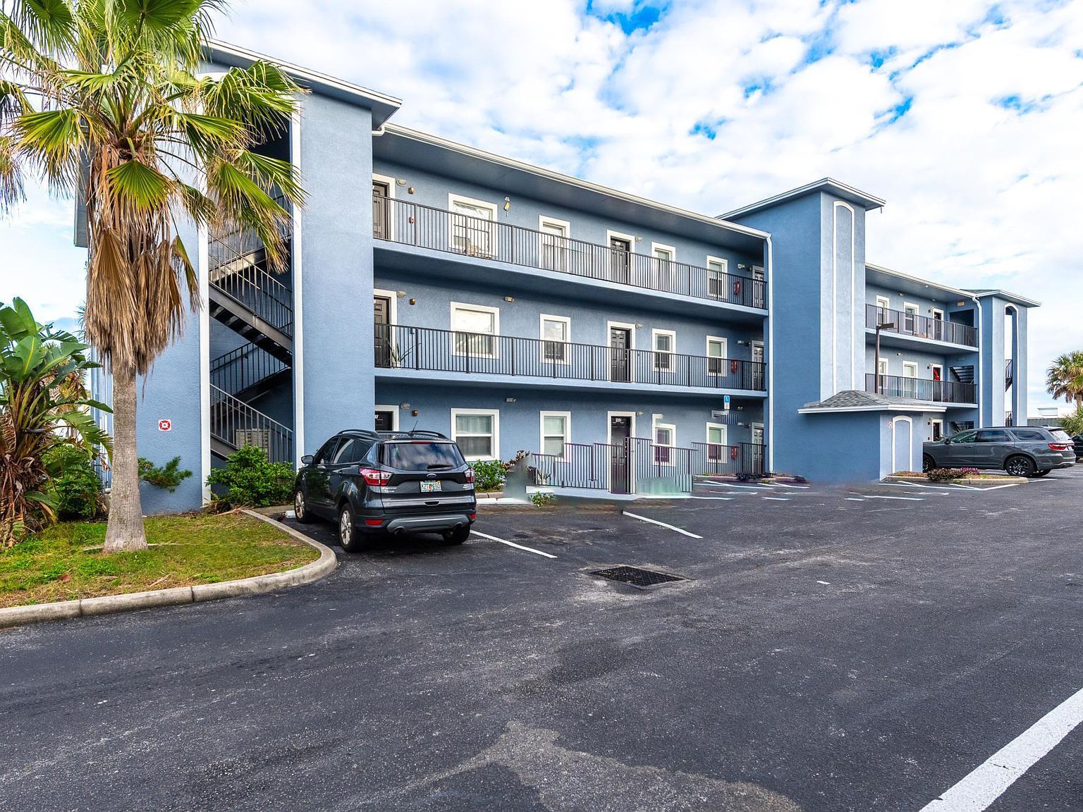 Blue multi-story apartment building with balconies, parked cars, and palm tree.