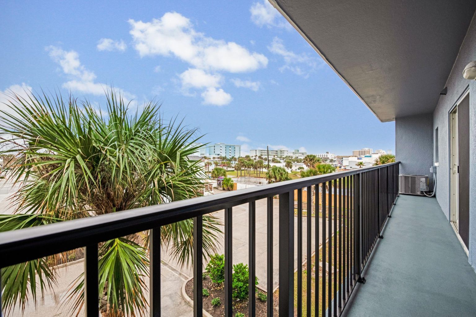 Balcony with black railing overlooks palm tree, parking lot, and city under cloudy sky.