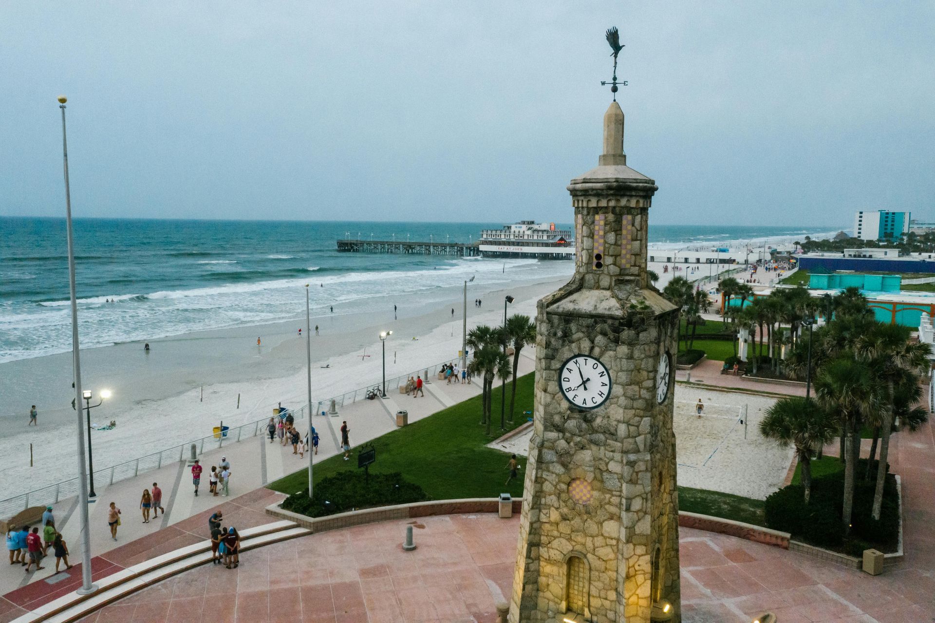 Stone clock tower on a beach boardwalk, ocean in background. People walk on the beach.
