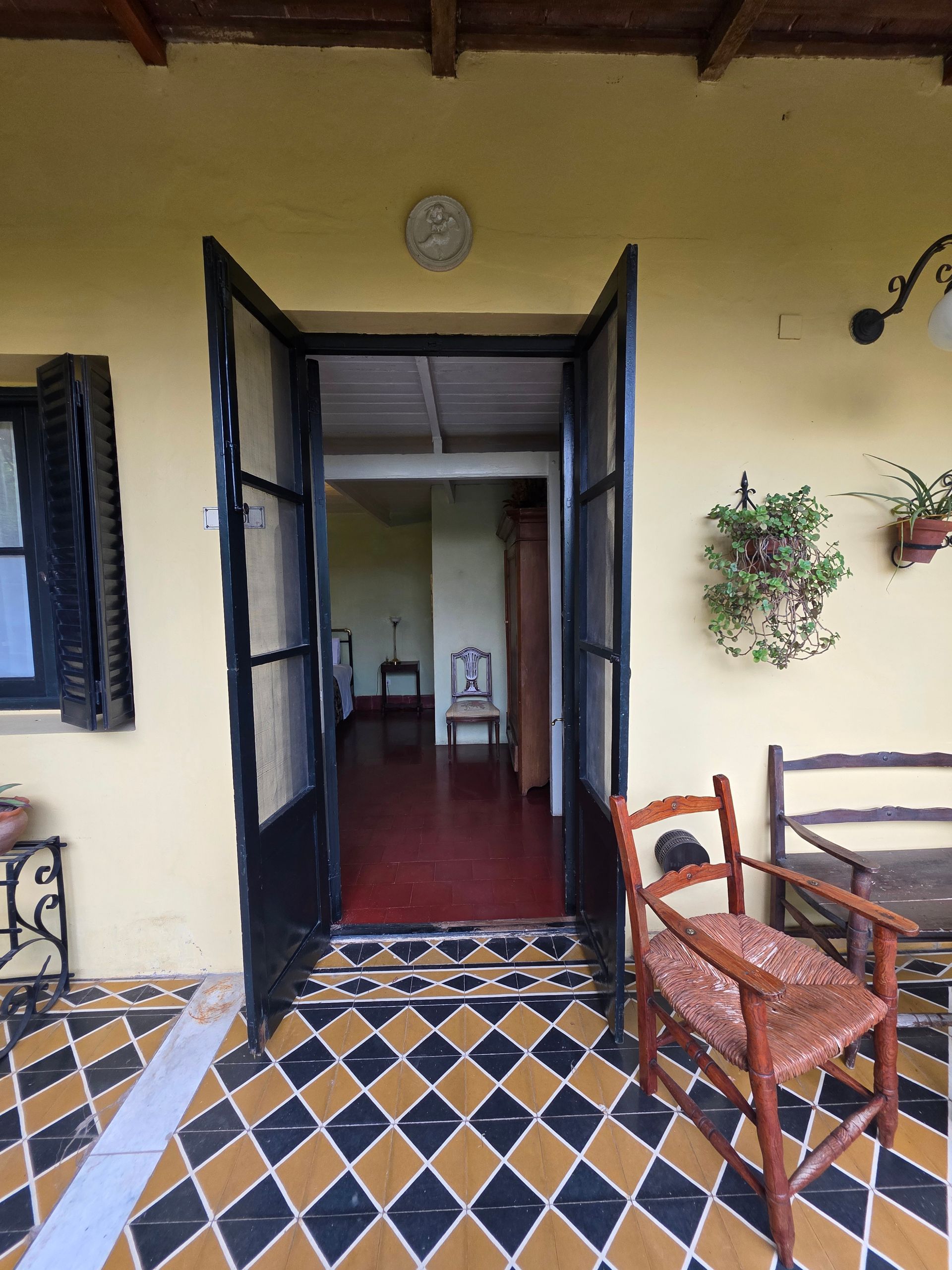 Unas puertas dobles negras abiertas revelan una habitación con suelo rojo. Un patio de baldosas tiene una silla y una planta cuelga de la pared.