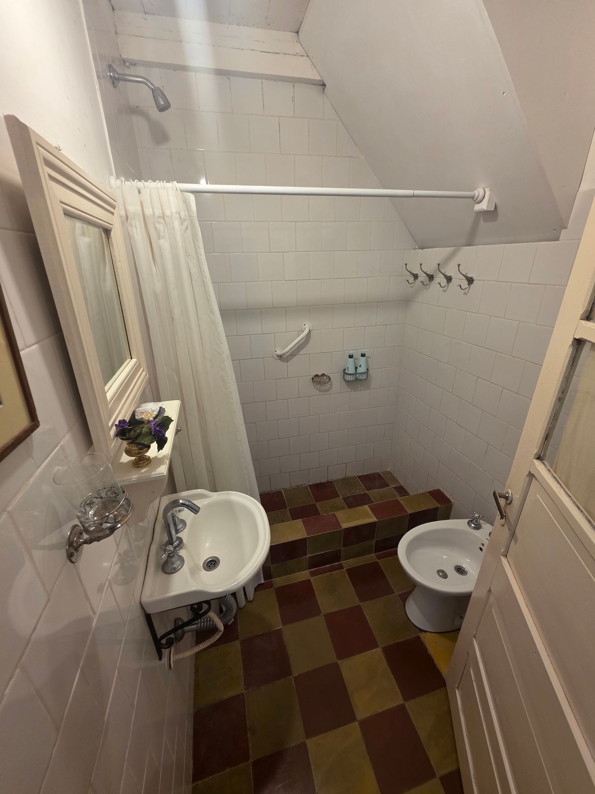 Baño pequeño con lavabo, espejo, ducha y bidé. Suelo de cuadros rojos y dorados, paredes de azulejos blancos.