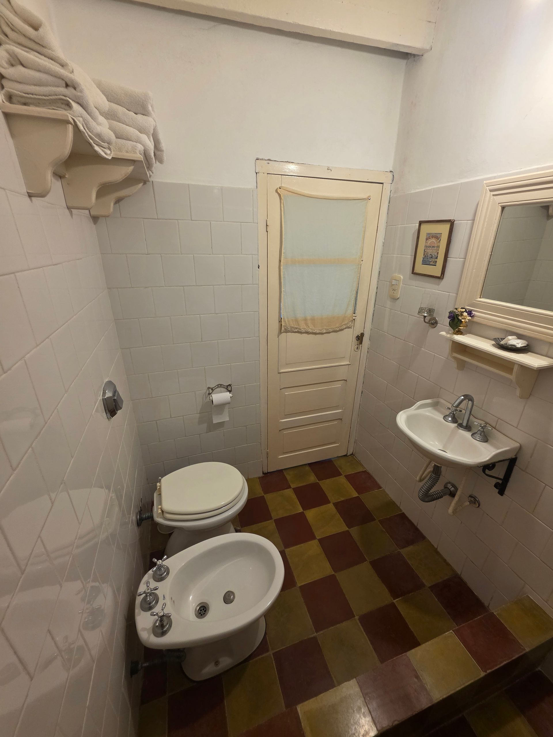 Pequeño baño con paredes blancas, suelo de baldosas, inodoro, bidet, lavabo y puerta abierta.
