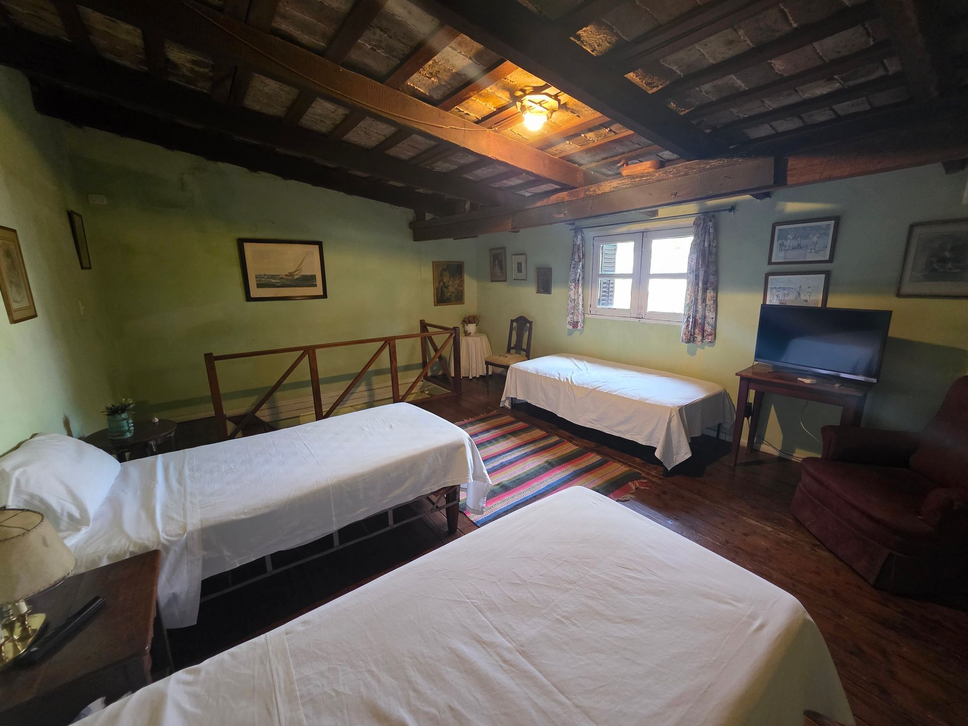 Dormitorio con tres camas individuales, paredes verdes, vigas de madera, pequeña ventana y obras de arte enmarcadas.