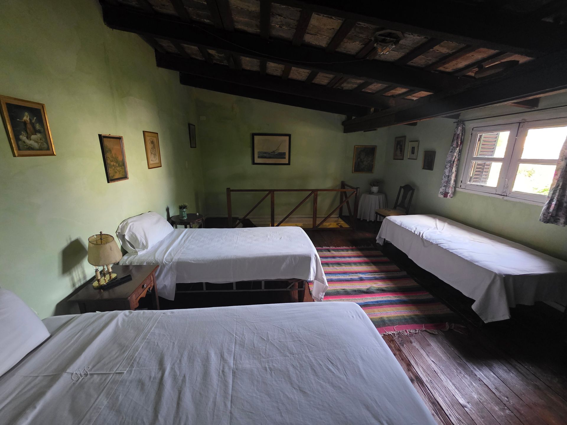 Dormitorio con tres camas, paredes verdes, vigas de madera en el techo, pequeñas obras de arte enmarcadas.