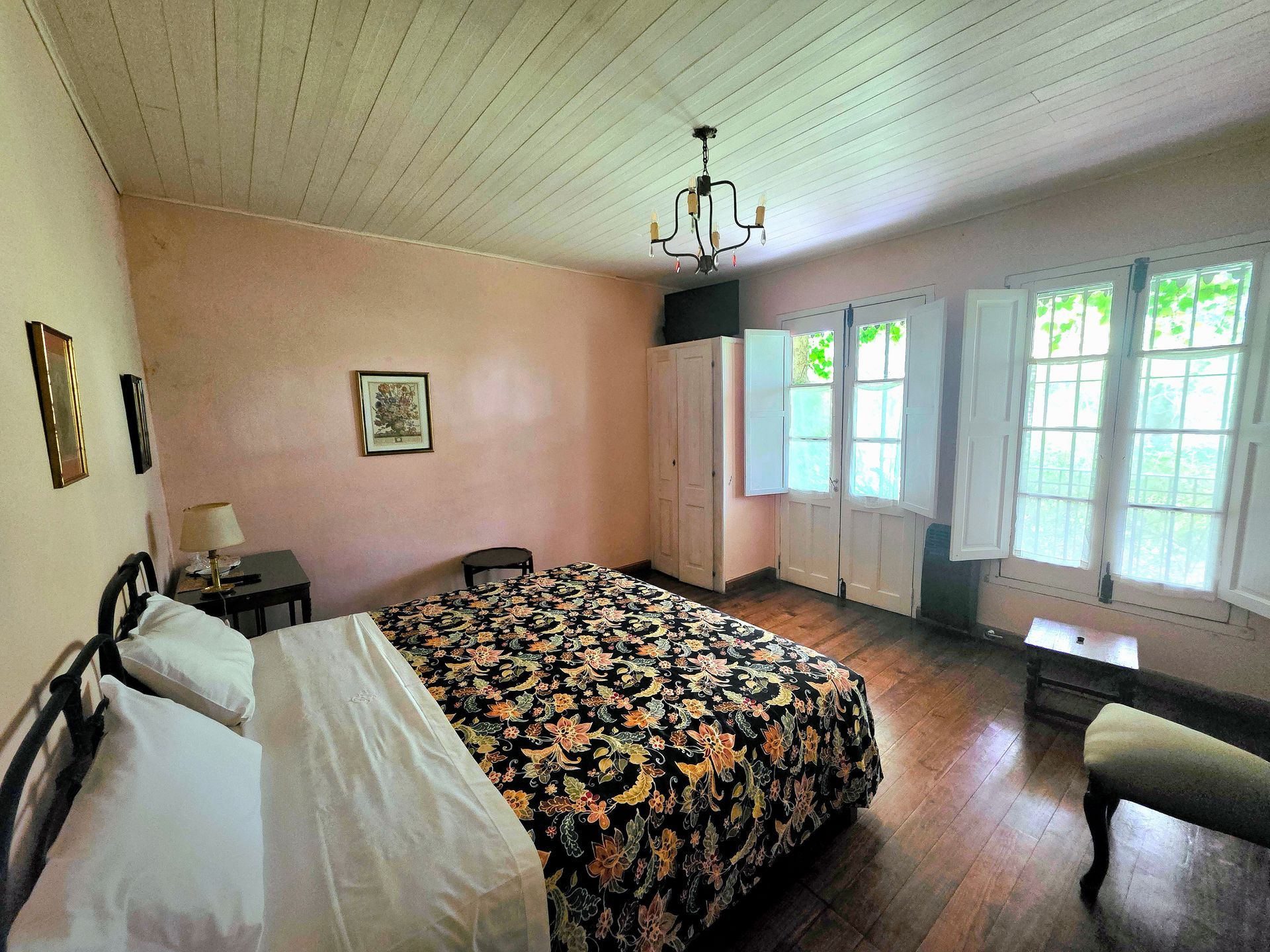 Dormitorio con colcha floral, suelo de madera y ventanas con contraventanas. Paredes rosas y lámpara de araña.