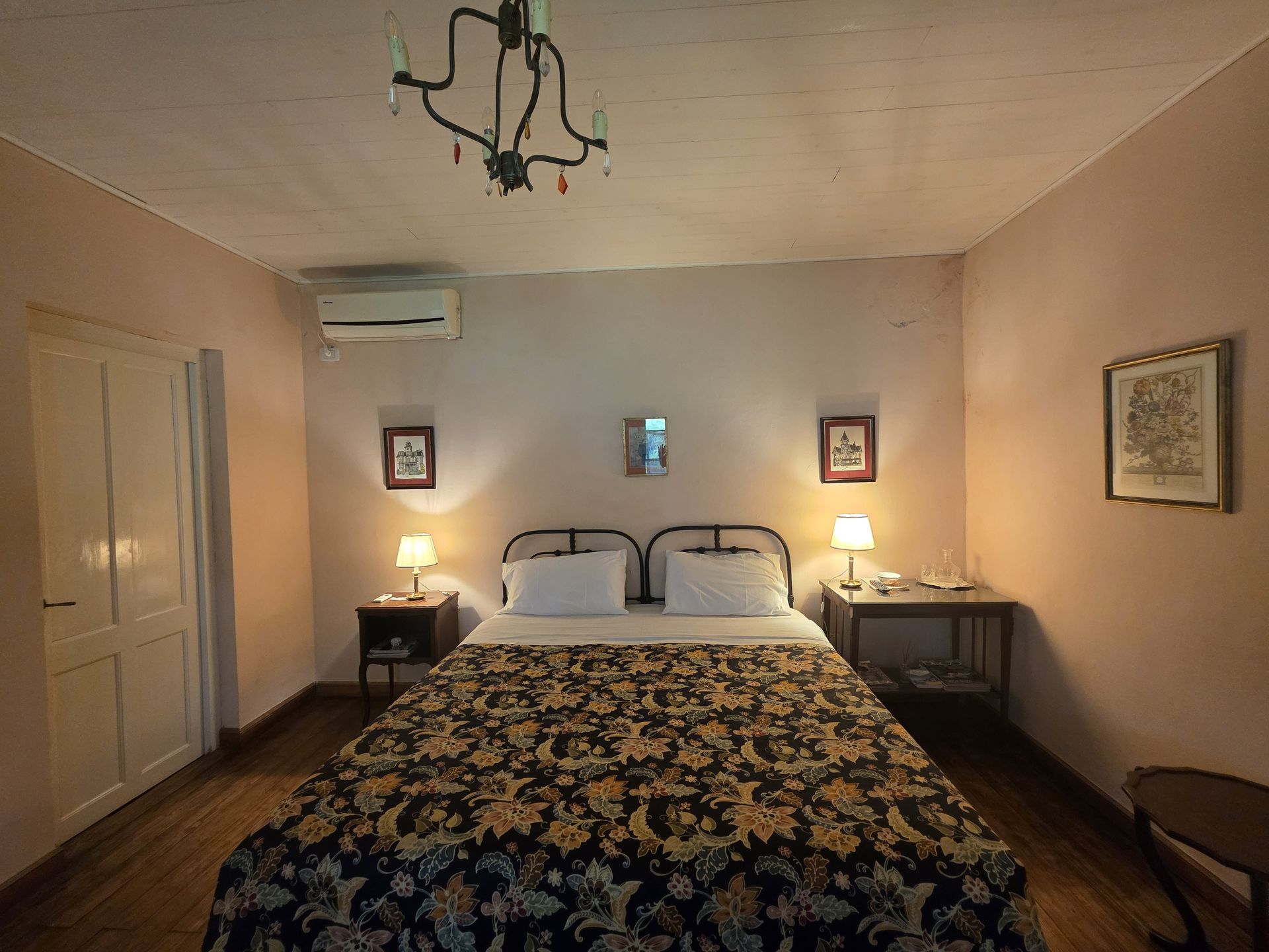 Dormitorio con cama, mesitas de noche, lámparas y obras de arte en paredes rosas. Sobre la cama hay una colcha oscura con estampado floral.