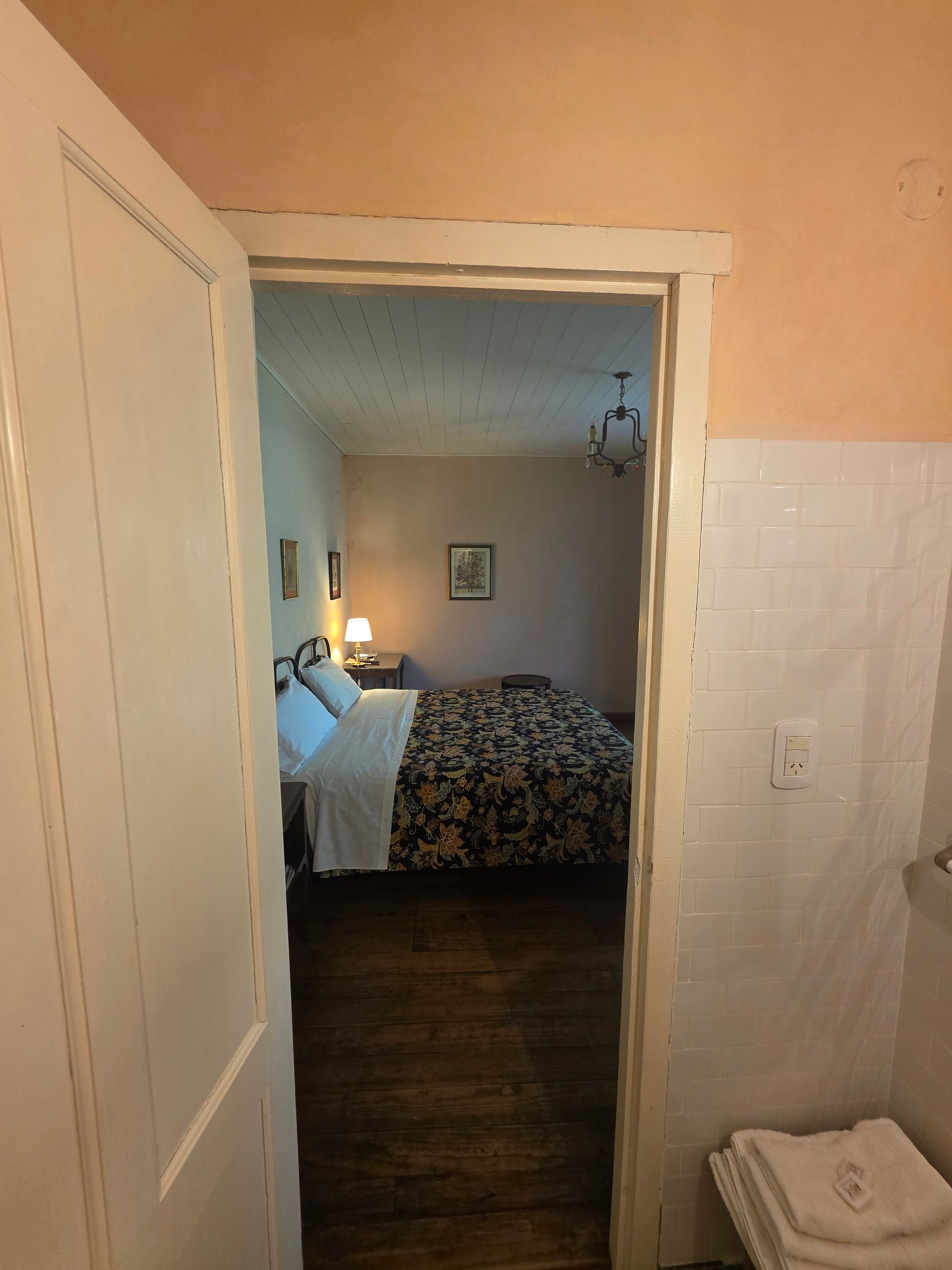 Puerta abierta a un dormitorio con cama. Edredón floral oscuro, suelo de madera y una pared en tonos cálidos.