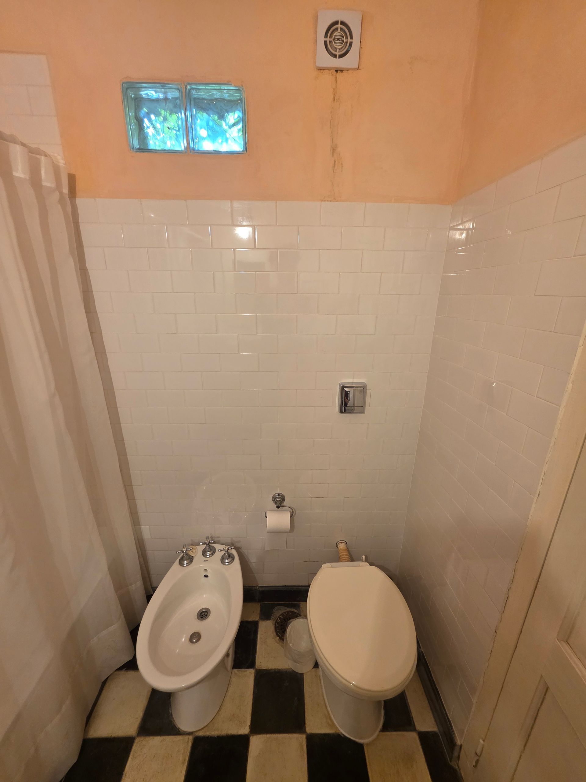 Baño con paredes de azulejos blancos, inodoro, bidé y suelo estampado en blanco y negro.