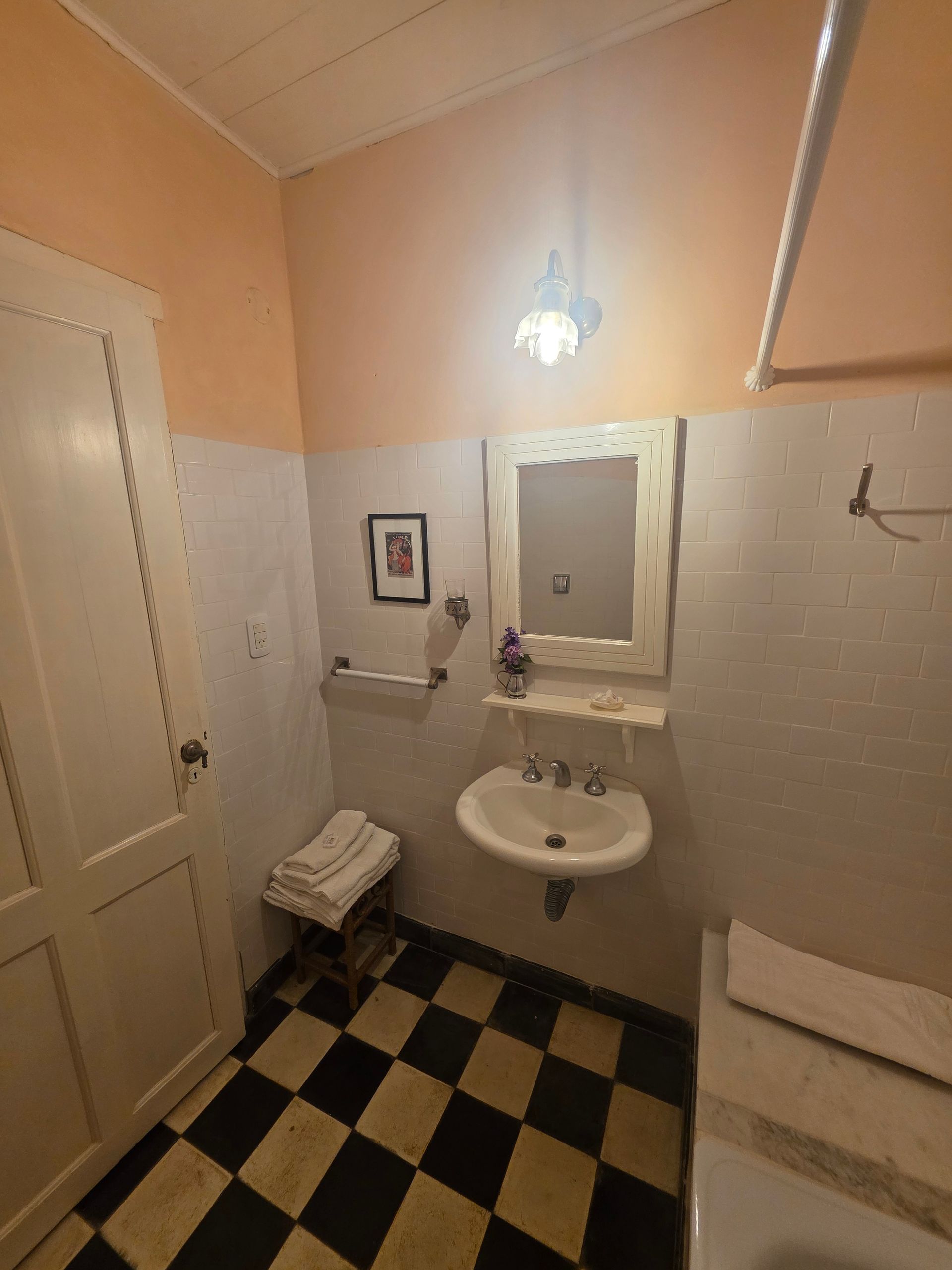 Pequeño baño con suelo a cuadros blancos y negros, azulejos blancos, paredes color melocotón, lavabo, espejo y toallas.