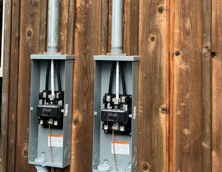 Two electrical meter boxes mounted on a weathered wood wall, with conduit pipes extending upwards.