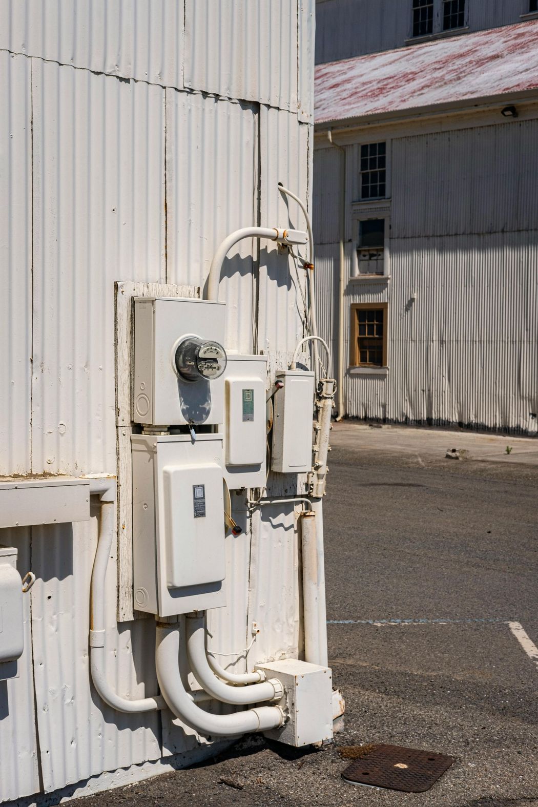 White electrical boxes and conduit on a corrugated white building exterior, with another building in the background.
