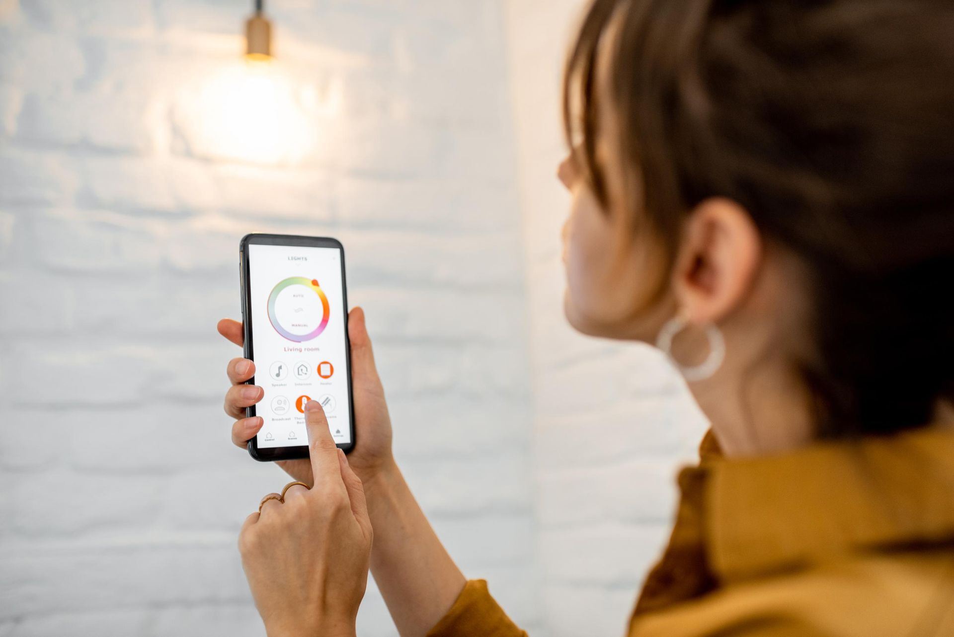Woman adjusting smart bulb color with phone, white brick wall background.