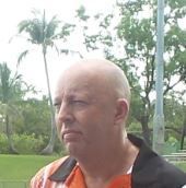 A bald man is standing in a park with trees in the background.