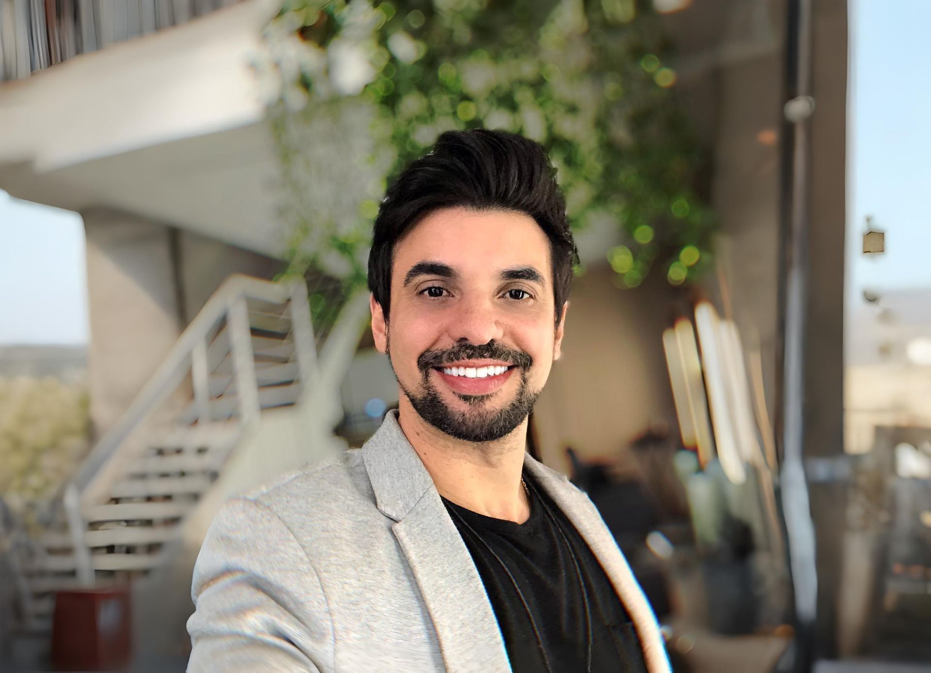 Homem sorridente com blazer cinza, camisa preta, ao ar livre, degraus e vegetação ao fundo.