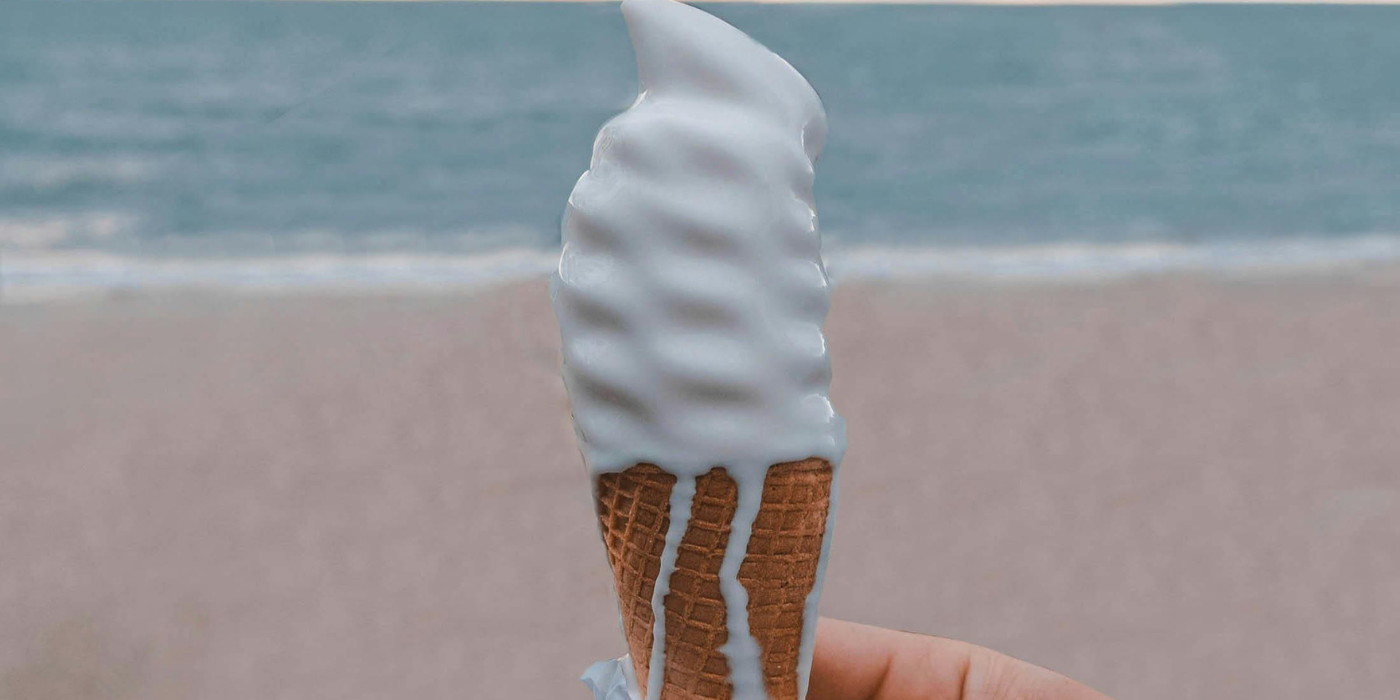 White soft-serve ice cream cone with a waffle cone, held against a blurred beach background