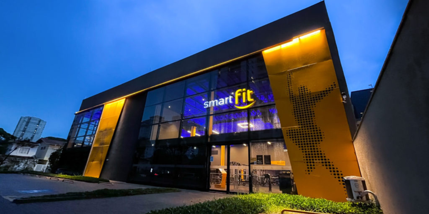 Exterior de uma academia Smart Fit ao entardecer, com janelas iluminadas em azul