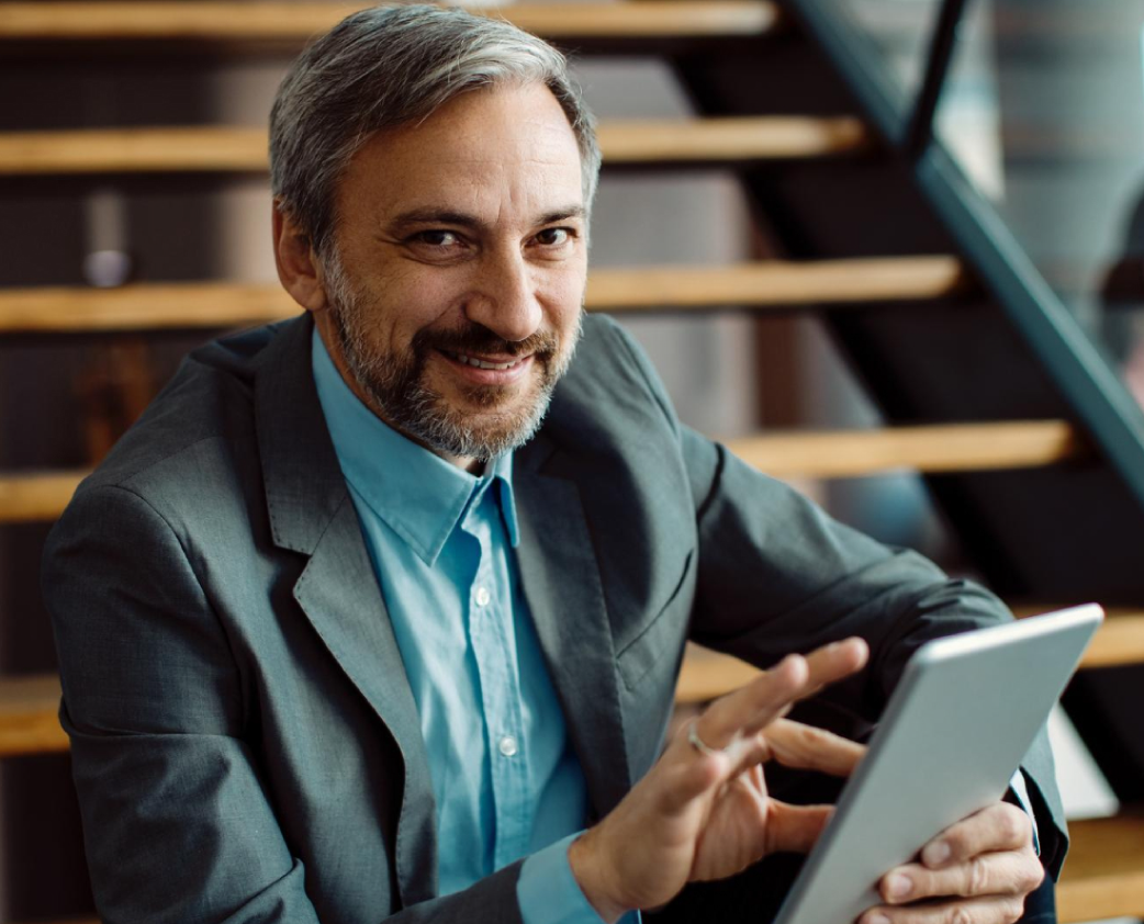 Homem com cabelos e barba grisalhos sorrindo, usando tablet em escadas de madeira.