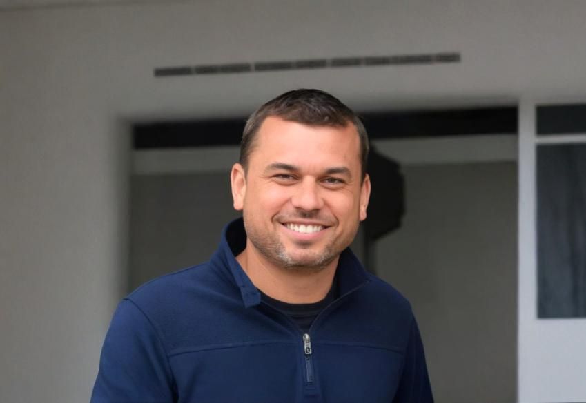 Homem sorridente com um suéter azul sobre um fundo branco.