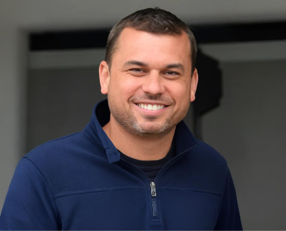 Homem com cabelo castanho curto sorrindo em um moletom azul com zíper ao ar livre.