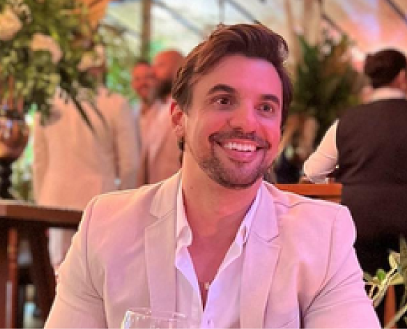 Homem de terno bege e camisa aberta sorri em uma festa com flores e pessoas ao fundo.