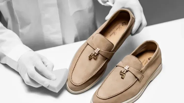 Mãos com luvas brancas apresentam dois mocassins de camurça bege sobre uma superfície branca.