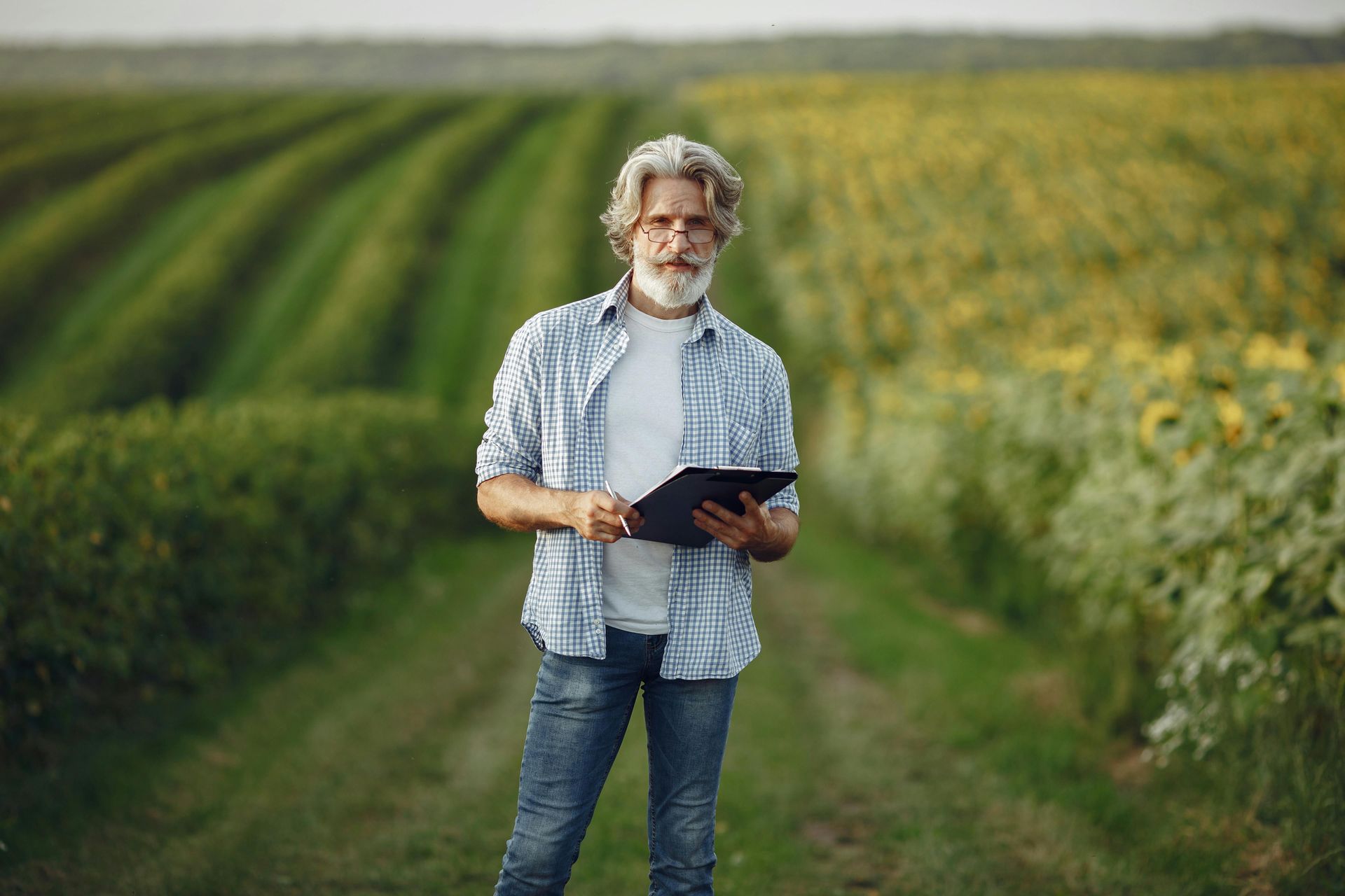 Homem segurando um tablet, em pé em um campo de plantação; vestindo uma camisa xadrez azul e calça 