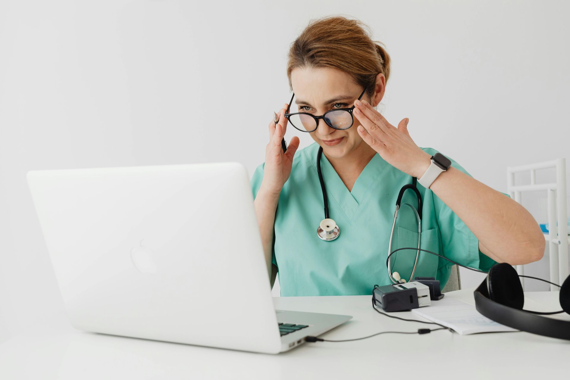Médica de uniforme cirúrgico olha para um laptop, ajustando os óculos em uma sala branca. 